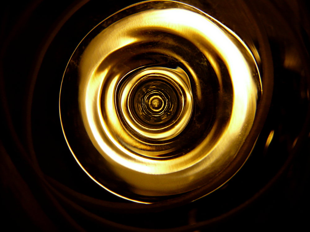 An abstract photograph of a wineglass held to a light, showing a golden, circular cascade of light and dark.