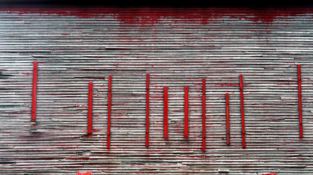 Numerous vertical red beams nailed to a dilapidated grey and white barn wall, seen as an experimental form of music notation.
