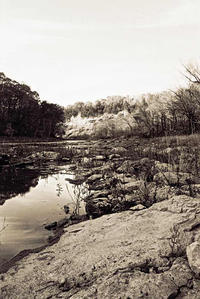 A monochrome rendering of the rocky shore of the Vermilion River, with darkened forests and a brilliantly lit bluff converging in the distance.
