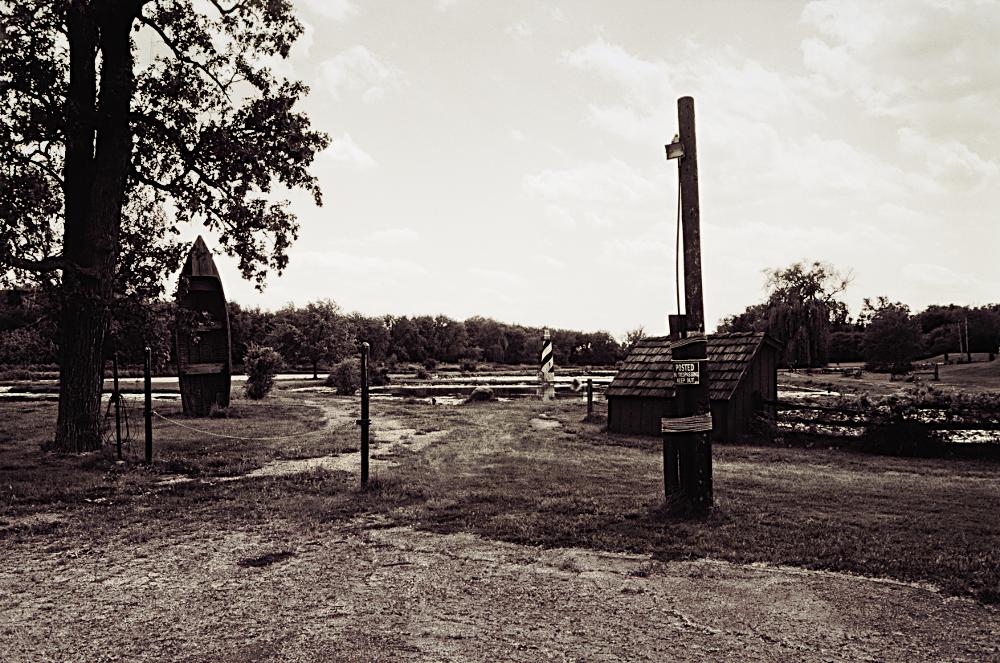 The edge of an abandoned parking lot and trail, with a distant lighthouse and decorative boat guarded by a no trespassing sign. Rendered in monochrome.