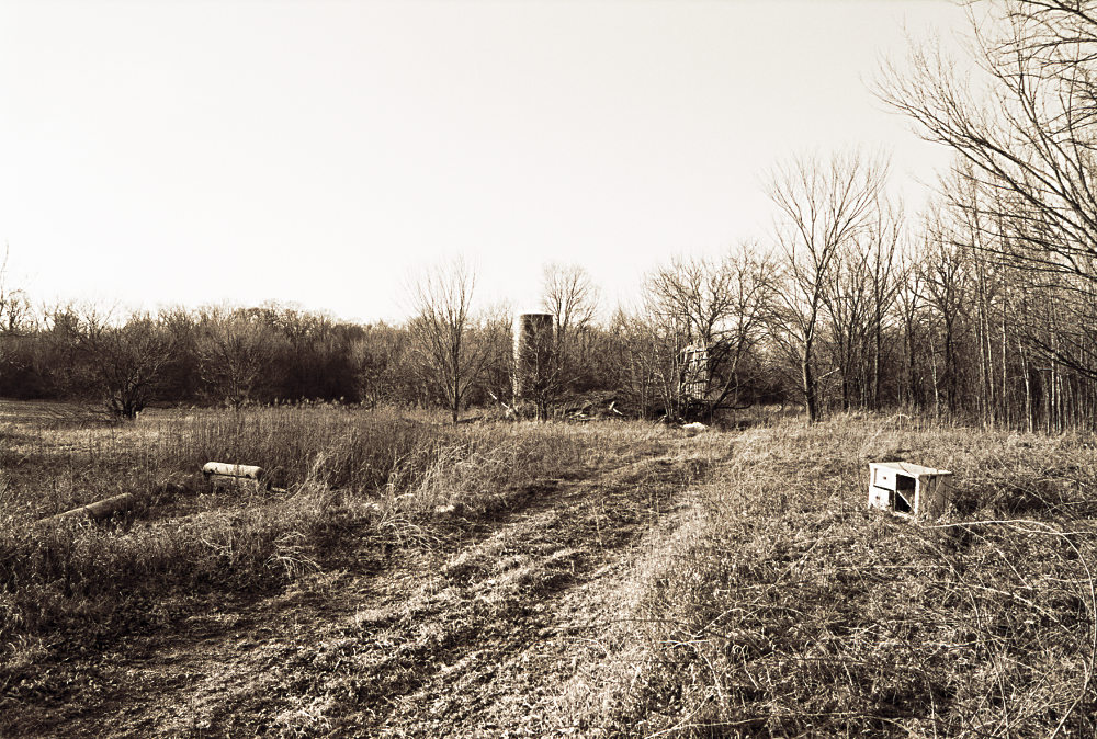 An abandoned plot of land with a fallen home and derelict silo in the distance, rendered in monochrome. Belongings are strewn about some tire-flattened grass.