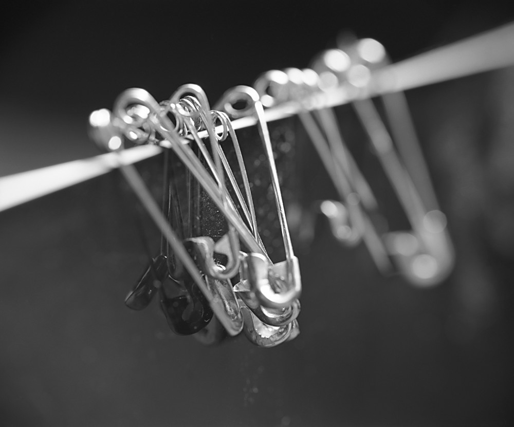 A monochrome rendering of safety pins suspended on a blurred, thin edge. The placement and varied focus conveys the motion of a galloping horse.
