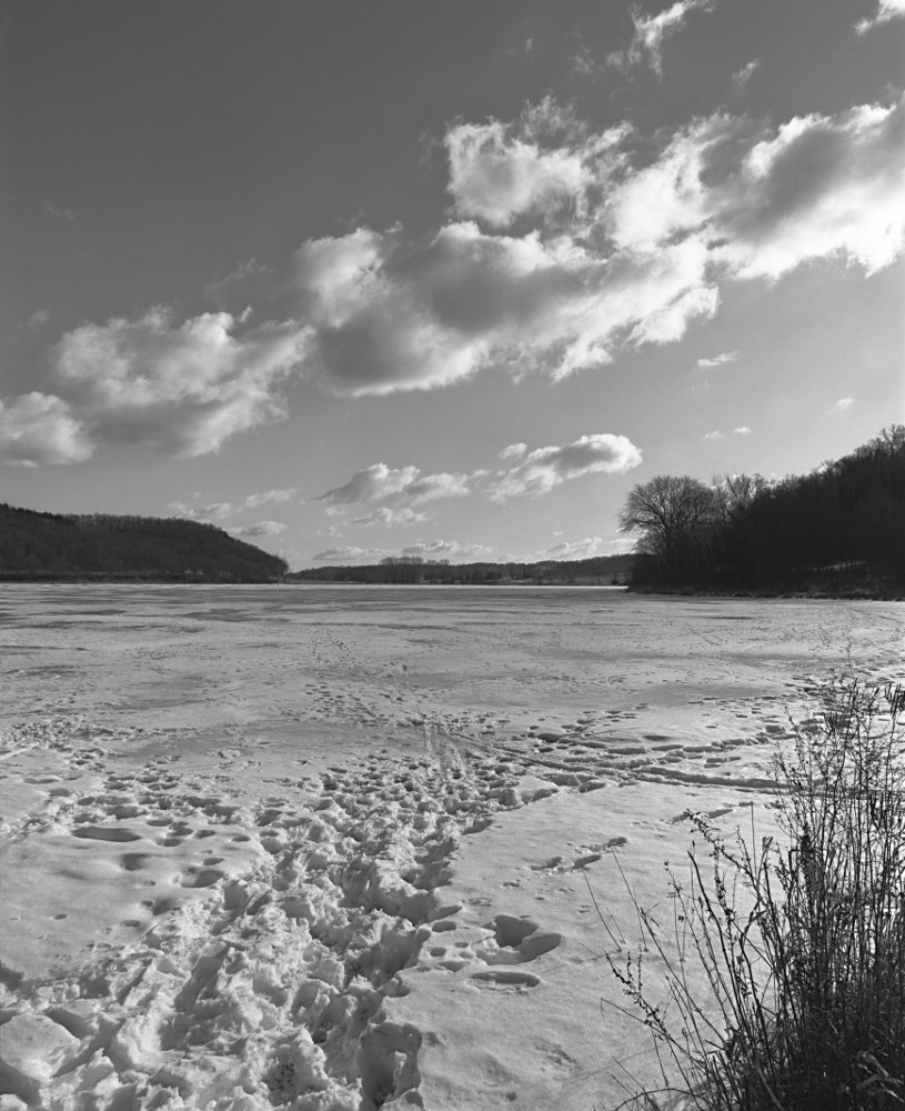 A lake edged by forests and covered in thick snow that is lined with adventurous footprints. Rendered in monochrome.